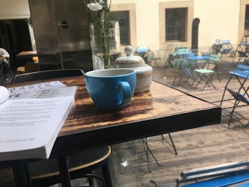 A cozy café setting with a blue cup on a wooden table, accompanied by a teapot and a book. In the background, there is an outdoor patio area with empty blue and green metal chairs and tables. A vase with white flowers adds a delicate touch to the table decor, while natural light streams in, giving a warm atmosphere.