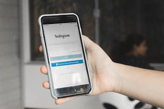 A hand holding a smartphone displaying the Instagram login screen. The phone is encased in a white cover, and the background appears blurred with the suggestion of an indoor setting featuring a chair and a person.