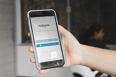 A hand holding a smartphone displaying the Instagram login screen. The phone is encased in a white cover, and the background appears blurred with the suggestion of an indoor setting featuring a chair and a person.