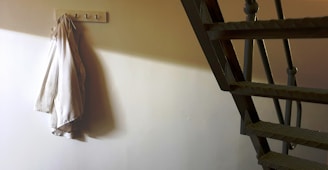 A fabric garment hangs on a wall hook in a dimly lit corner with shadowed stairs leading upwards, emphasizing a minimalist setting.