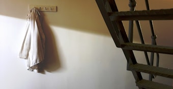 A fabric garment hangs on a wall hook in a dimly lit corner with shadowed stairs leading upwards, emphasizing a minimalist setting.