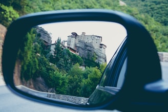 A scenic view of a stone monastery perched on a rocky cliff surrounded by lush greenery, reflected in the side mirror of a car.