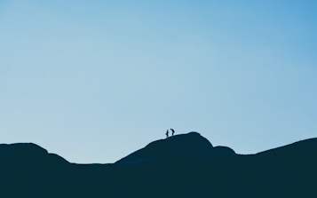 Silhouettes of two people stand atop a rocky hill against a clear sky, creating a sense of adventure and exploration.