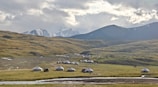 A rural landscape features a cluster of traditional yurts scattered across a vast, grassy plain. In the background, snow-capped mountains rise under a partly cloudy sky. A small stream meanders through the foreground, adding to the serene and picturesque setting.