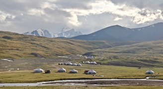 A rural landscape features a cluster of traditional yurts scattered across a vast, grassy plain. In the background, snow-capped mountains rise under a partly cloudy sky. A small stream meanders through the foreground, adding to the serene and picturesque setting.