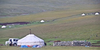 A vast, grassy landscape with several yurts scattered across the field. A white vehicle is parked near a prominent yurt, and there is a stack of stones nearby. Small figures and horses can be seen in the distance, indicating a nomadic setting.