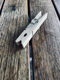A wooden clothespin rests on a textured wooden surface, showcasing its simple metal spring mechanism. The wood has a natural finish with visible grain patterns, adding a rustic feel.