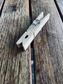 A wooden clothespin rests on a textured wooden surface, showcasing its simple metal spring mechanism. The wood has a natural finish with visible grain patterns, adding a rustic feel.