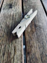 A wooden clothespin rests on a textured wooden surface, showcasing its simple metal spring mechanism. The wood has a natural finish with visible grain patterns, adding a rustic feel.