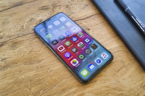 A smartphone with a vibrant screen displaying various app icons lies on a wooden surface. Next to the phone, a black notebook and a pen are placed.
