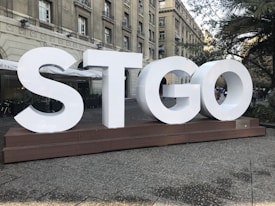 Large white letters spelling 'STGO' are displayed on a wooden platform. The structure is situated in an outdoor area with a historical building in the background. Trees and plants are part of the setting, and people walk around in the distance.