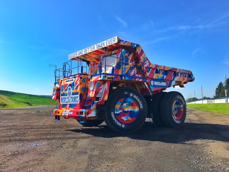 A large dump truck with a colorful, vibrant paint job, featuring various patterns and logos including 'World of Tanks.' It is parked on a dirt path under a clear blue sky, with patches of grass and trees in the background. The truck has oversized wheels and a sign that reads 'Bigger and Better Than Ever!'