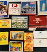 A collection of matchbox covers displayed on a flat surface. They feature a variety of designs and branding, including hotels and different artistic motifs. The matchboxes have vivid illustrations and various texts in multiple languages, indicating places like Brazil, India, and the USA.