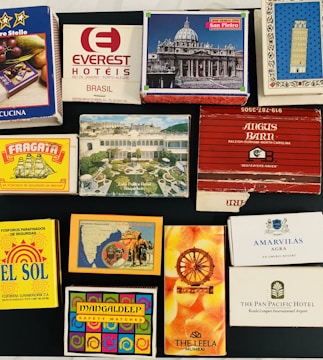 A collection of matchbox covers displayed on a flat surface. They feature a variety of designs and branding, including hotels and different artistic motifs. The matchboxes have vivid illustrations and various texts in multiple languages, indicating places like Brazil, India, and the USA.