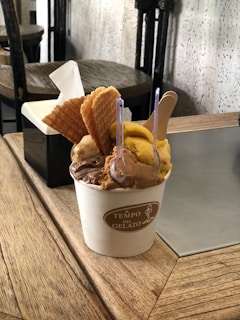 A cup of gelato with multiple flavors is presented on a wooden table. The gelato is topped with several pieces of waffle cone and served with two small clear spoons. A tissue box is visible in the background, and the setting includes a rustic wooden decor.