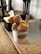 A cup of gelato with multiple flavors is presented on a wooden table. The gelato is topped with several pieces of waffle cone and served with two small clear spoons. A tissue box is visible in the background, and the setting includes a rustic wooden decor.
