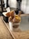 A cup of gelato with multiple flavors is presented on a wooden table. The gelato is topped with several pieces of waffle cone and served with two small clear spoons. A tissue box is visible in the background, and the setting includes a rustic wooden decor.