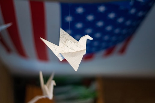 An origami paper crane made from a piece of lined paper with writing, suspended in the air. The background features a blurred American flag with red and white stripes and white stars on a blue field.
