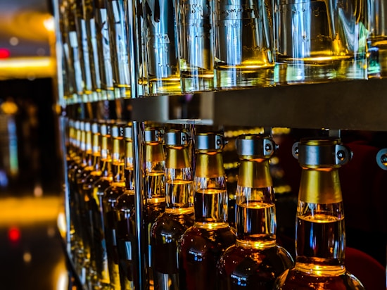 Rows of glass bottles filled with a golden liquid are aligned on shelves in a dimly lit setting. The bottles have metallic caps and emit a warm amber glow, reflecting the light in the room.