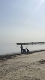 A person is standing on a sandy beach, holding a fishing rod and fishing by a calm body of water. Beside them is a scooter parked on the sand. The sky is clear, and the overall atmosphere is peaceful and serene.