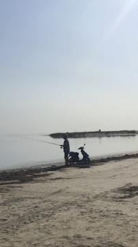 A person is standing on a sandy beach, holding a fishing rod and fishing by a calm body of water. Beside them is a scooter parked on the sand. The sky is clear, and the overall atmosphere is peaceful and serene.