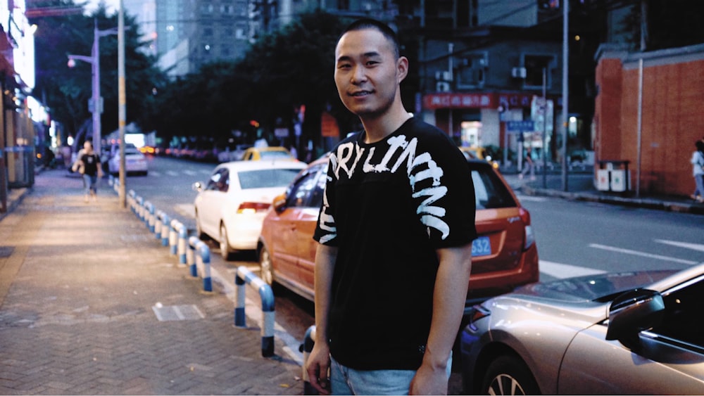 man wearing black and white shirt standing beside parked vehicles beside the street