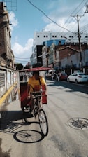 A bicycle taxi is being pedaled down a city street. The rider wears a yellow shirt and brown pants. The street is lined with various buildings and cars are visible in the background. Overhead, the sky is mostly clear with some clouds. The sun casts shadows from the bicycle onto the pavement.