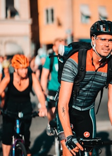 A group of cyclists is riding on a street, with one cyclist in the foreground wearing a striped jersey, helmet, glasses, and backpack. The background shows blurred buildings and other cyclists. The scene takes place in a sunny urban environment.