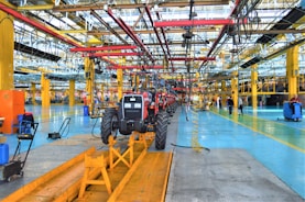 A large indoor factory setting with a partially assembled tractor situated prominently in the center. The factory is equipped with various machinery and equipment, including overhead conveyor systems with suspended components. The environment is spacious, well-lit, featuring expansive use of steel beams and painted floors. Workers are visible in the background engaged in various tasks.