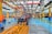 A large indoor factory setting with a partially assembled tractor situated prominently in the center. The factory is equipped with various machinery and equipment, including overhead conveyor systems with suspended components. The environment is spacious, well-lit, featuring expansive use of steel beams and painted floors. Workers are visible in the background engaged in various tasks.