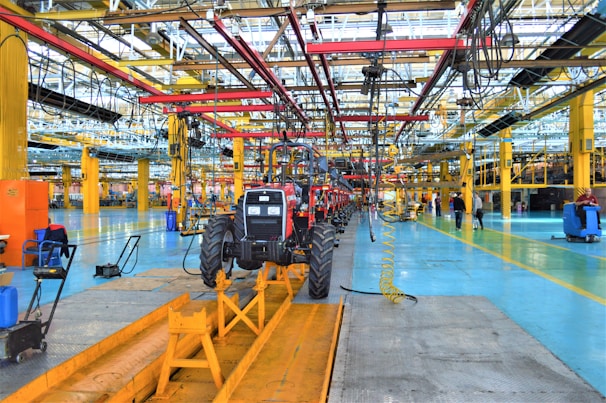 A large indoor factory setting with a partially assembled tractor situated prominently in the center. The factory is equipped with various machinery and equipment, including overhead conveyor systems with suspended components. The environment is spacious, well-lit, featuring expansive use of steel beams and painted floors. Workers are visible in the background engaged in various tasks.