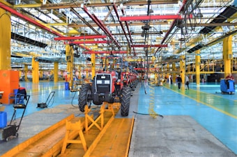 A large indoor factory setting with a partially assembled tractor situated prominently in the center. The factory is equipped with various machinery and equipment, including overhead conveyor systems with suspended components. The environment is spacious, well-lit, featuring expansive use of steel beams and painted floors. Workers are visible in the background engaged in various tasks.