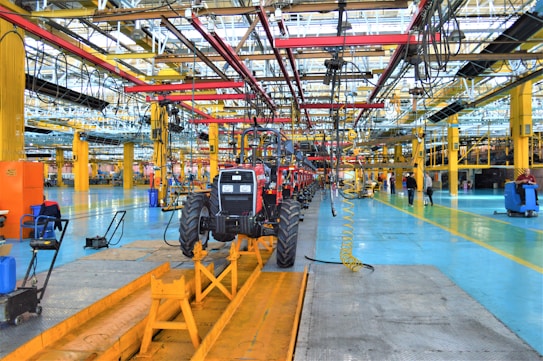 A large indoor factory setting with a partially assembled tractor situated prominently in the center. The factory is equipped with various machinery and equipment, including overhead conveyor systems with suspended components. The environment is spacious, well-lit, featuring expansive use of steel beams and painted floors. Workers are visible in the background engaged in various tasks.