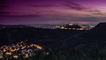A nighttime landscape featuring a brightly lit village nestled within a valley. In the distance, a solitary illuminated building sits atop a hill, surrounded by trees. The expansive sky is tinged with deep purples and subtle oranges as the sun sets or rises, casting a serene and enchanting atmosphere.