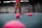 The image depicts a tennis court with a person standing, holding a tennis racket. There are numerous pink tennis balls scattered across the blue court surface. The person's attire includes pink shorts and shoes, matching the color of the tennis balls. The background shows a tennis academy with some branding visible on the walls.