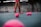 The image depicts a tennis court with a person standing, holding a tennis racket. There are numerous pink tennis balls scattered across the blue court surface. The person's attire includes pink shorts and shoes, matching the color of the tennis balls. The background shows a tennis academy with some branding visible on the walls.