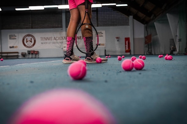 The image depicts a tennis court with a person standing, holding a tennis racket. There are numerous pink tennis balls scattered across the blue court surface. The person's attire includes pink shorts and shoes, matching the color of the tennis balls. The background shows a tennis academy with some branding visible on the walls.