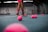 The image depicts a tennis court with a person standing, holding a tennis racket. There are numerous pink tennis balls scattered across the blue court surface. The person's attire includes pink shorts and shoes, matching the color of the tennis balls. The background shows a tennis academy with some branding visible on the walls.