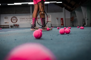 The image depicts a tennis court with a person standing, holding a tennis racket. There are numerous pink tennis balls scattered across the blue court surface. The person's attire includes pink shorts and shoes, matching the color of the tennis balls. The background shows a tennis academy with some branding visible on the walls.