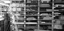A cluttered workshop or storage space filled with shelves stacked with various materials and objects. There are numerous containers, wooden planks, and assorted construction materials organized in a haphazard manner. Labels and numbers are visible on the shelves, indicating an attempt at categorization.