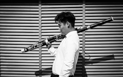 A person holding a bassoon over their shoulder, standing in front of a slatted wall. The individual is wearing a white shirt and appears to be ready for a performance or rehearsal. The image is in black and white, emphasizing the contrast and texture of the scene.