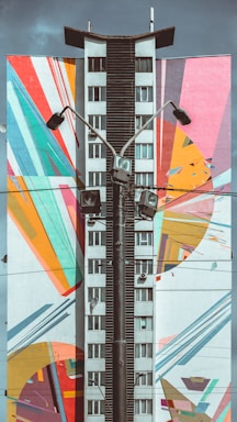 A tall building facade features a vibrant mural with geometric and abstract patterns in multiple colors. The center of the building is painted in a neutral tone, contrasting with the colorful artwork that includes arcs, stripes, and angular shapes. Several streetlights and a tangle of wires run vertically in front of the building.
