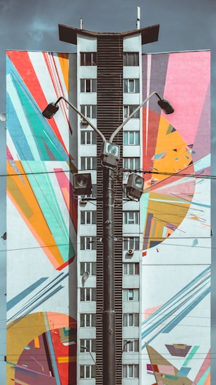 A tall building facade features a vibrant mural with geometric and abstract patterns in multiple colors. The center of the building is painted in a neutral tone, contrasting with the colorful artwork that includes arcs, stripes, and angular shapes. Several streetlights and a tangle of wires run vertically in front of the building.