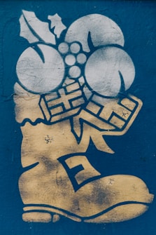 A graffiti-style artwork featuring a stylized boot in gold with decorations such as holly leaves, three round objects resembling berries or ornaments, and two gift box shapes. The background is a textured blue surface.