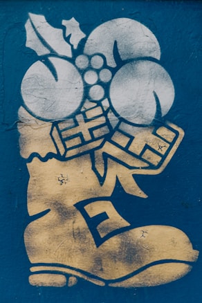 A graffiti-style artwork featuring a stylized boot in gold with decorations such as holly leaves, three round objects resembling berries or ornaments, and two gift box shapes. The background is a textured blue surface.