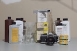 A vintage Nikon camera is surrounded by various photographic developing supplies, including canisters of chemical solutions, measuring beakers, and boxes of film. Several bottles from brands like Ilford and Kodak are present, as well as stainless steel tanks and beakers of different sizes placed on a flat surface.