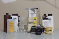 A vintage Nikon camera is surrounded by various photographic developing supplies, including canisters of chemical solutions, measuring beakers, and boxes of film. Several bottles from brands like Ilford and Kodak are present, as well as stainless steel tanks and beakers of different sizes placed on a flat surface.