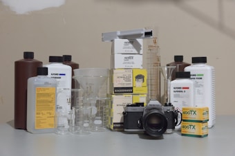 A vintage Nikon camera is surrounded by various photographic developing supplies, including canisters of chemical solutions, measuring beakers, and boxes of film. Several bottles from brands like Ilford and Kodak are present, as well as stainless steel tanks and beakers of different sizes placed on a flat surface.