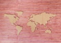 A wooden map of the world with continents cut out and placed on a woven bamboo background. The map features simplified shapes of all major continents with distinct outlines. The background consists of horizontal bamboo slats with varying shades of red and brown, creating a textured appearance.