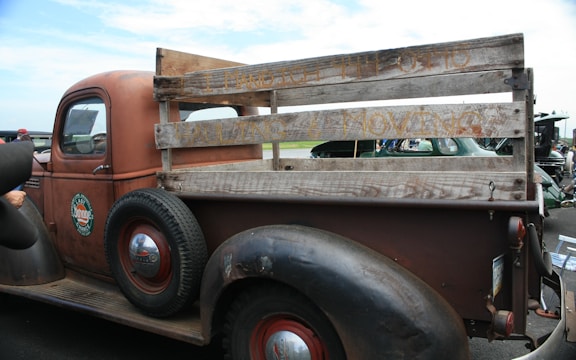 A vintage brown pickup truck with a wooden plank bed, showcasing old-fashioned design. The truck features a weathered look with visible rust and text on the wood panels. It has round, black tires with red rims and a classic 'Esso' logo on the driver's door.